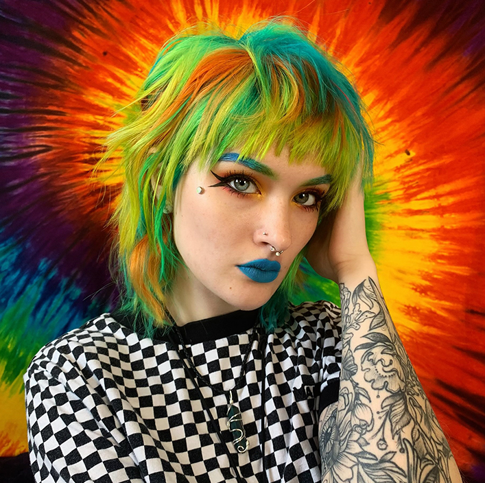 girl with green, yellow and orange hair in black and white checked shirt in front of tie-dye wall